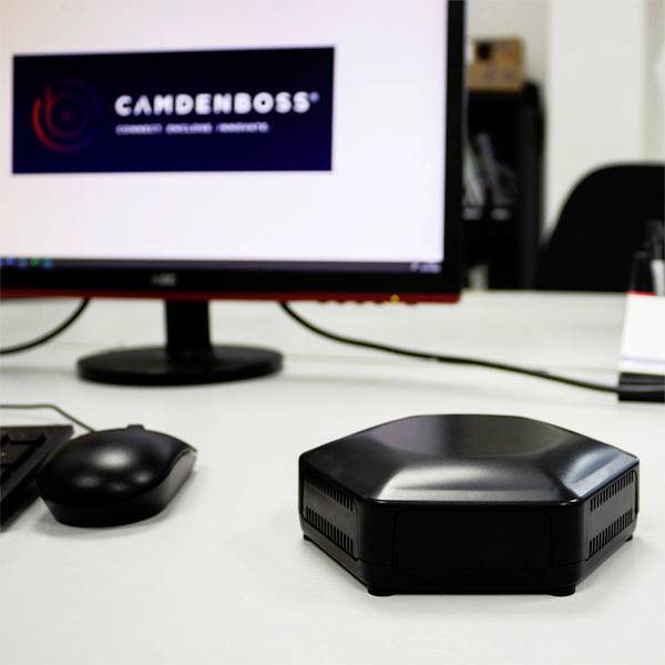 A black, hexagonal device sits on a desk in front of a computer monitor displaying the 'CAMDENBOSS' logo.