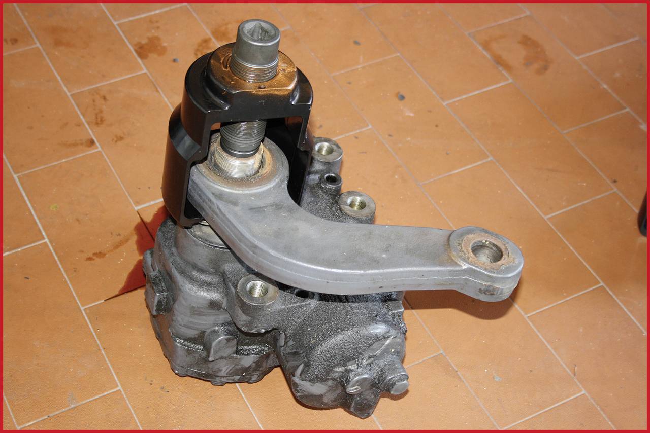 A mechanical car part is lying on an orange tiled floor. It is made of metal with a lever and several screw fastenings.