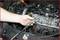 A mechanic is holding a spark plug in front of an open engine block, surrounded by various car parts and tools.