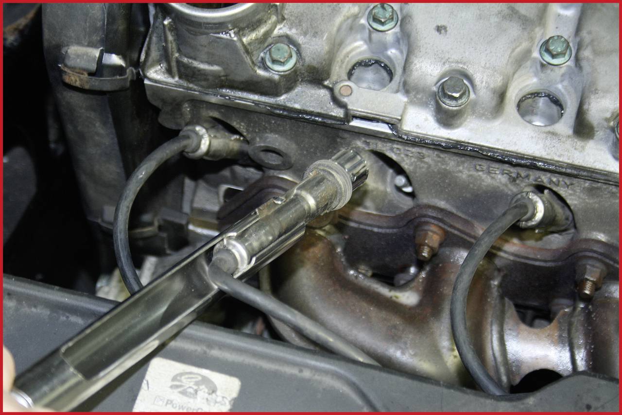 A spark plug is screwed into a car engine's spark plug hole, surrounded by cables and engine components.