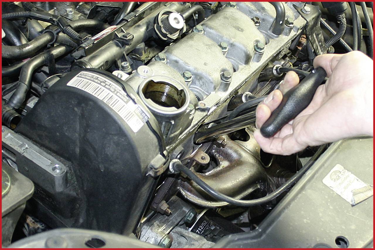 A hand is holding a screwdriver and working on an engine inside a car. The engine is partially open.