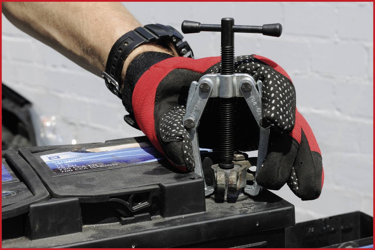 A mechanic carefully removes a battery terminal clamp using a puller while wearing gloves.