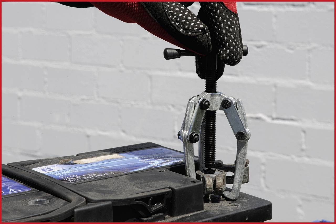 A gloved hand uses a puller to remove a clamp from a car battery. A brick wall is in the background.