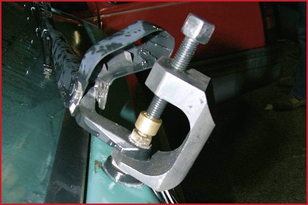Metal tool secured with a G-clamp attached to a green vehicle component against a red background.