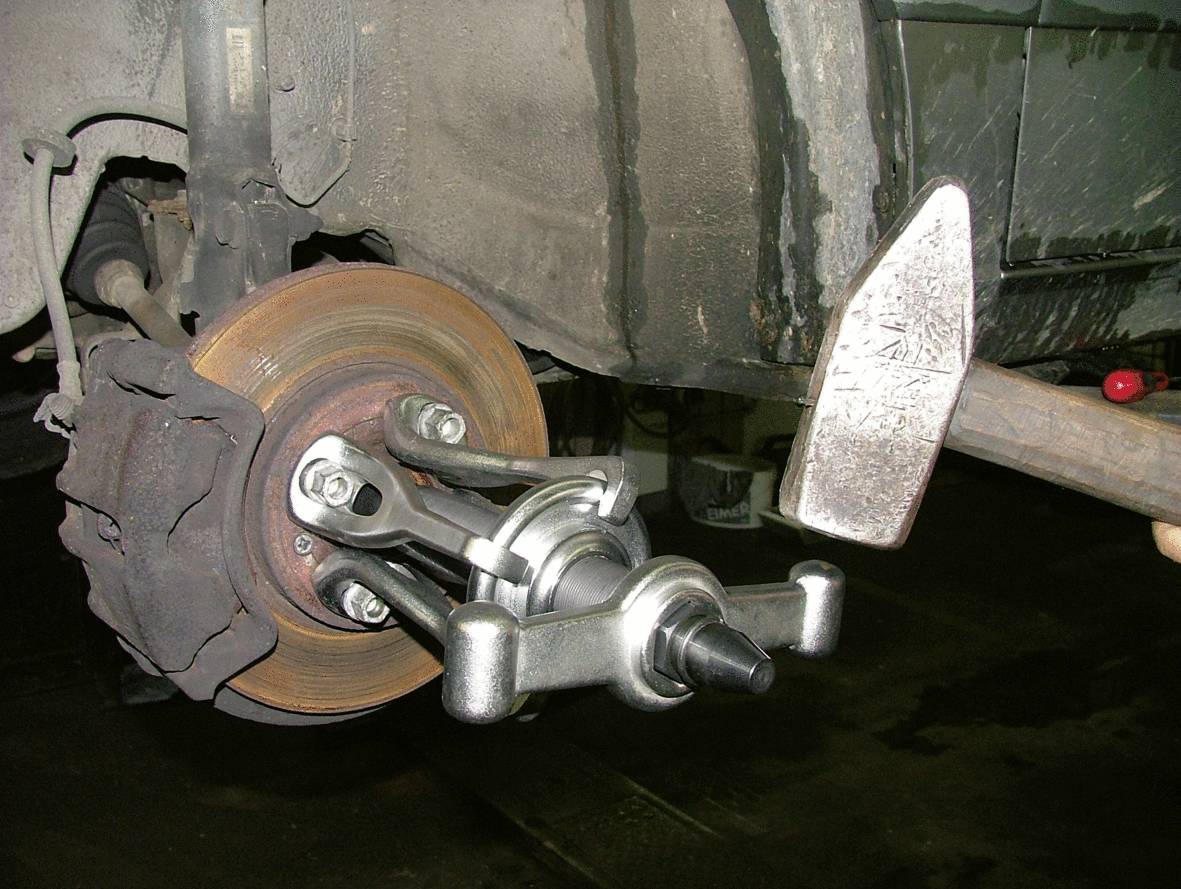 A wheel bearing hub is being removed from a vehicle's axle using a specialised puller. The brake disc is visible in the background.