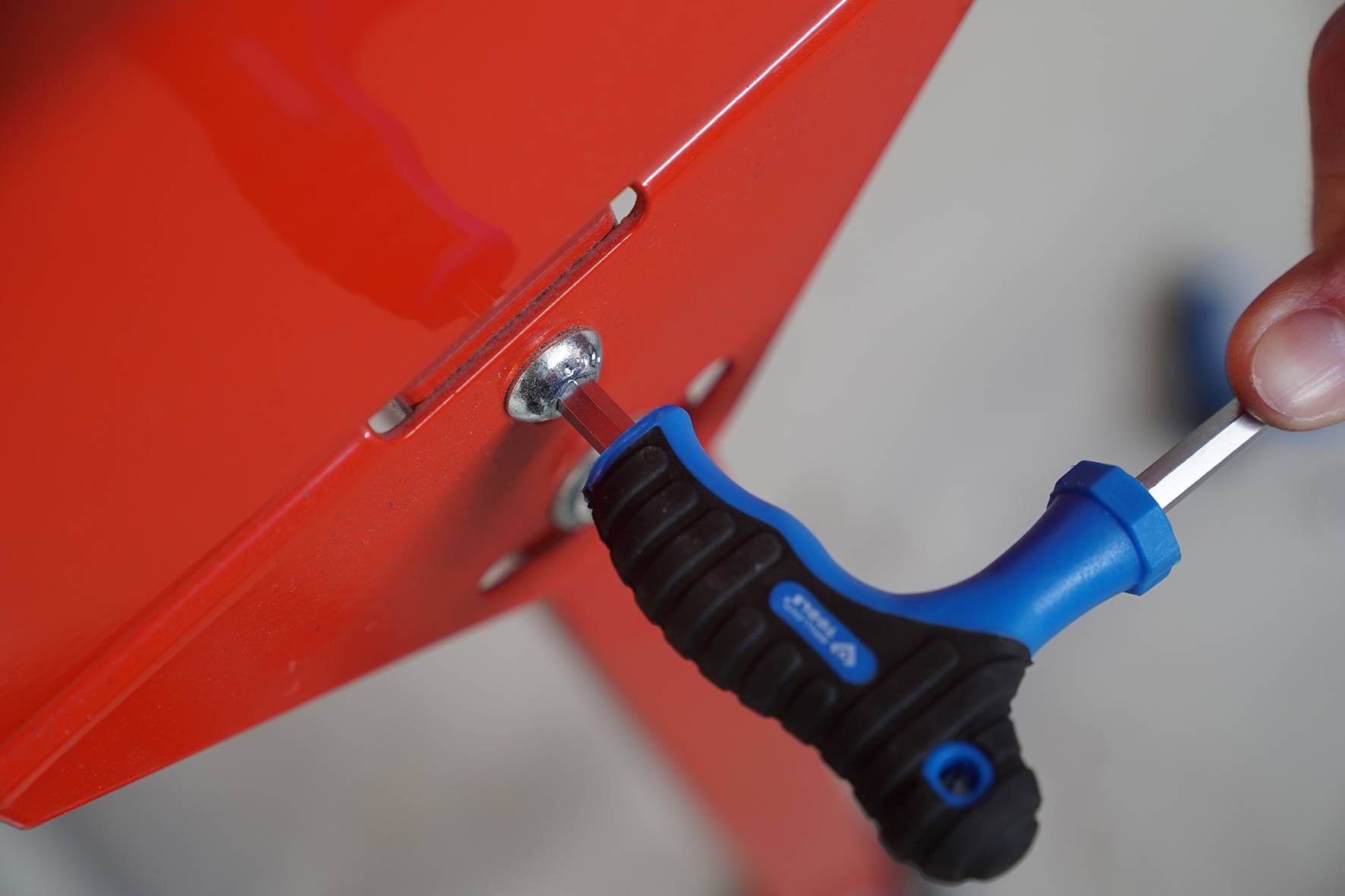 A person is using a blue and black screwdriver to tighten a screw on a red metal part.