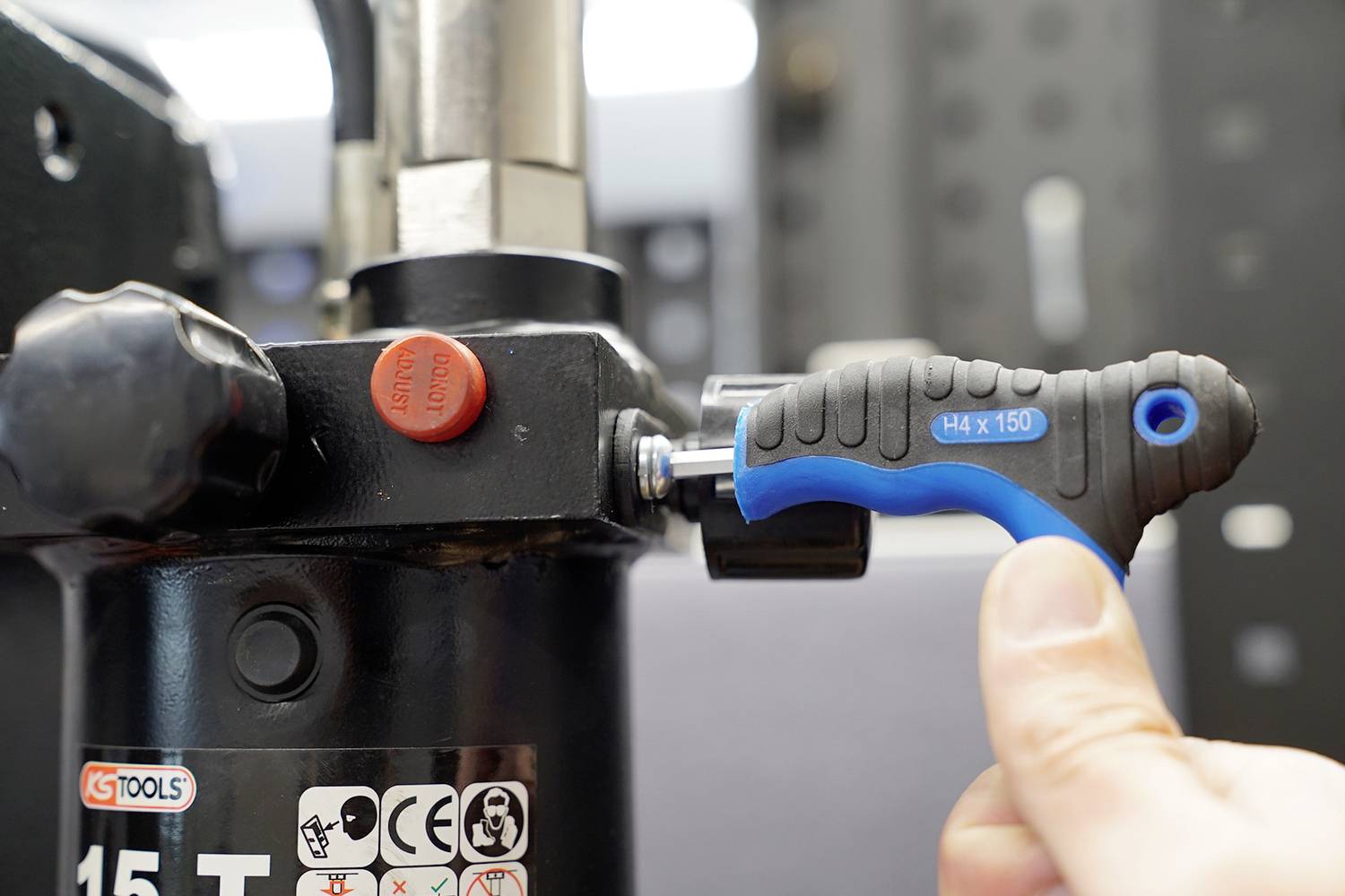 A hand is turning a blue screwdriver on a device with a black lever. The device shows printed markings and is in a workshop environment.