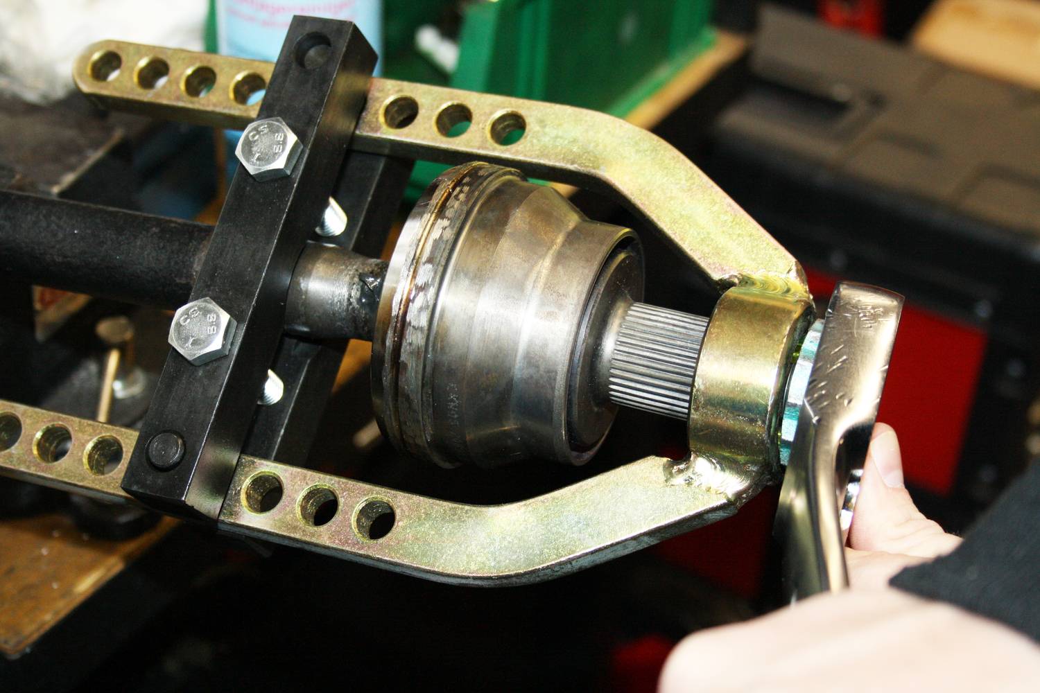 A hand is using a tool to secure or work on a metal part, presumably a vehicle component, surrounded by workshop items.