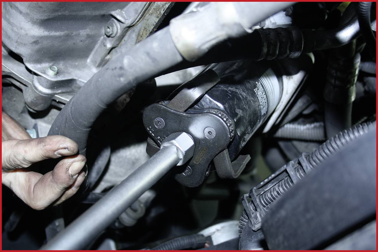 Mechanic's hand using a tool to remove an oil filter in the engine bay. Environment shows cables and hoses.