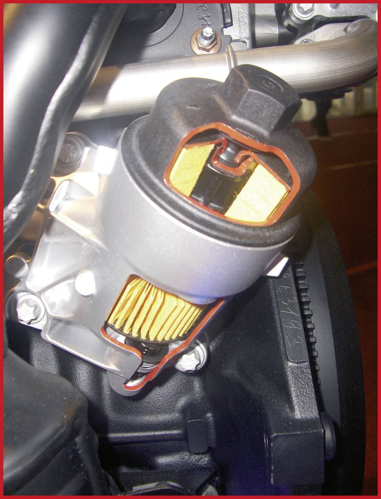 A silver oil filter with a yellow filter element is mounted in a metallic holder on a black engine.
