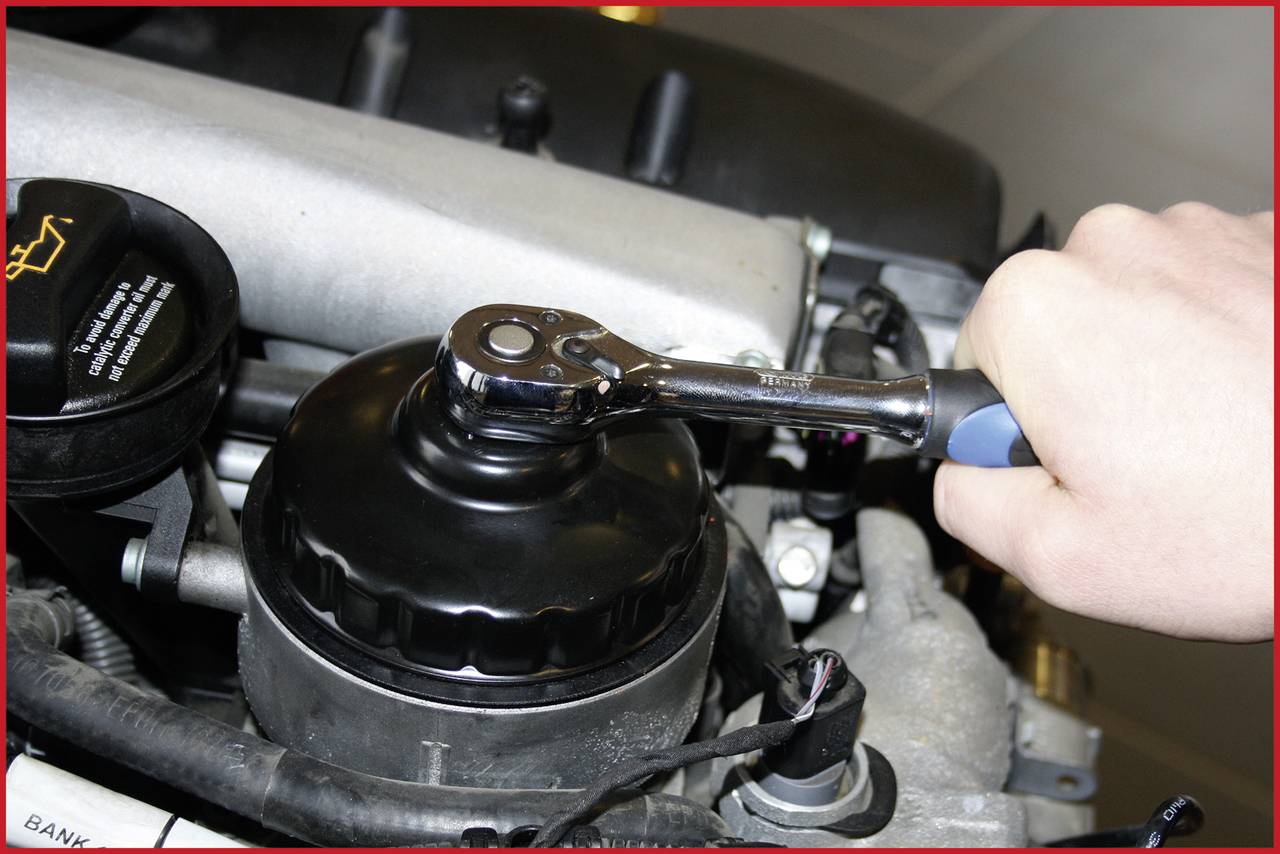 A hand is tightening a motor cover with a ratchet spanner. An engine is visible in the background.