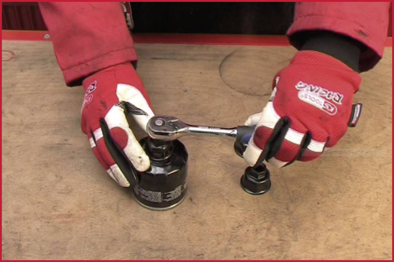 A person wearing red gloves is using a ratchet spanner to remove an oil filter on a wooden table.