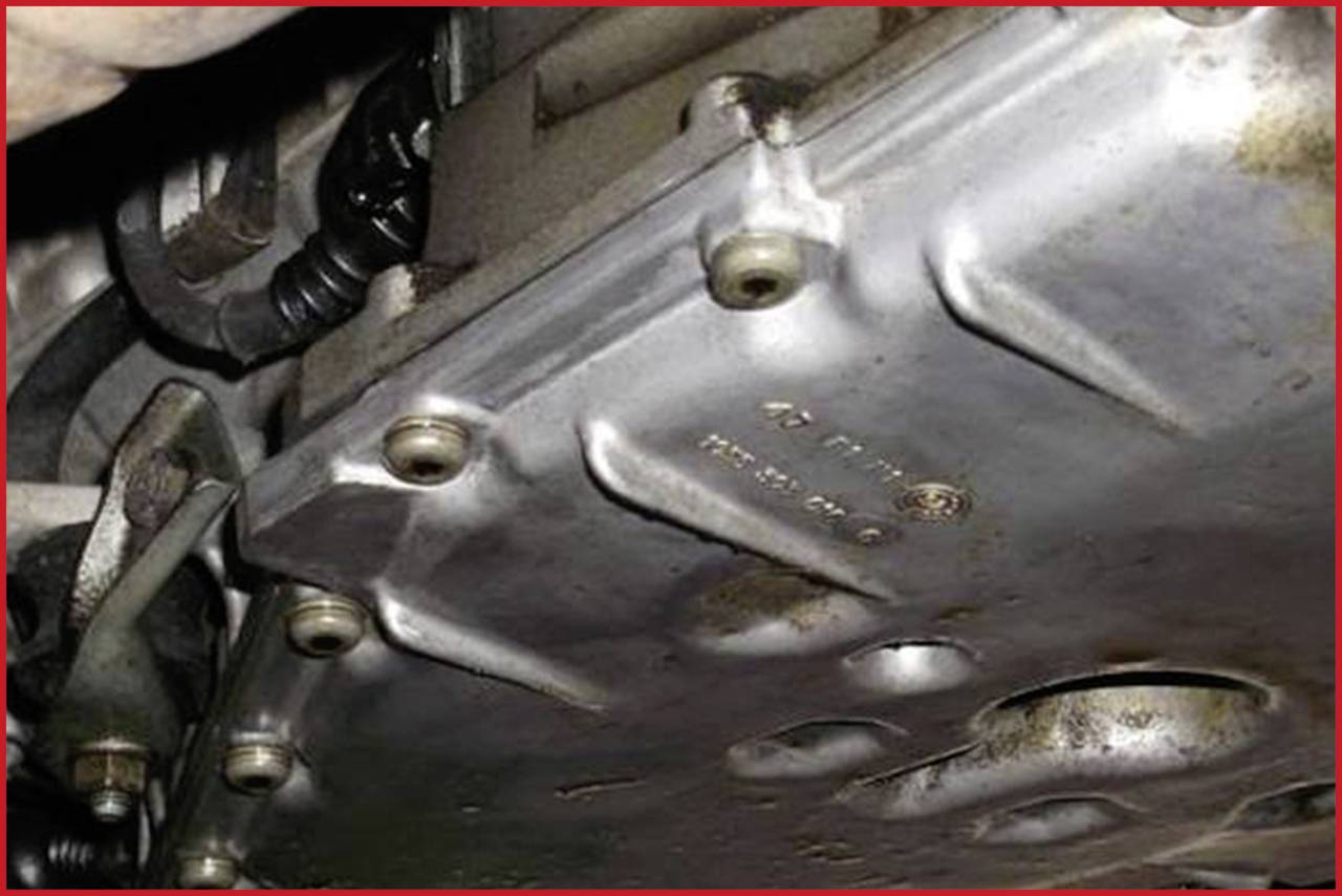 A gearbox housing with several screws and light rust. Structures and cables are visible in a vehicle undercarriage.