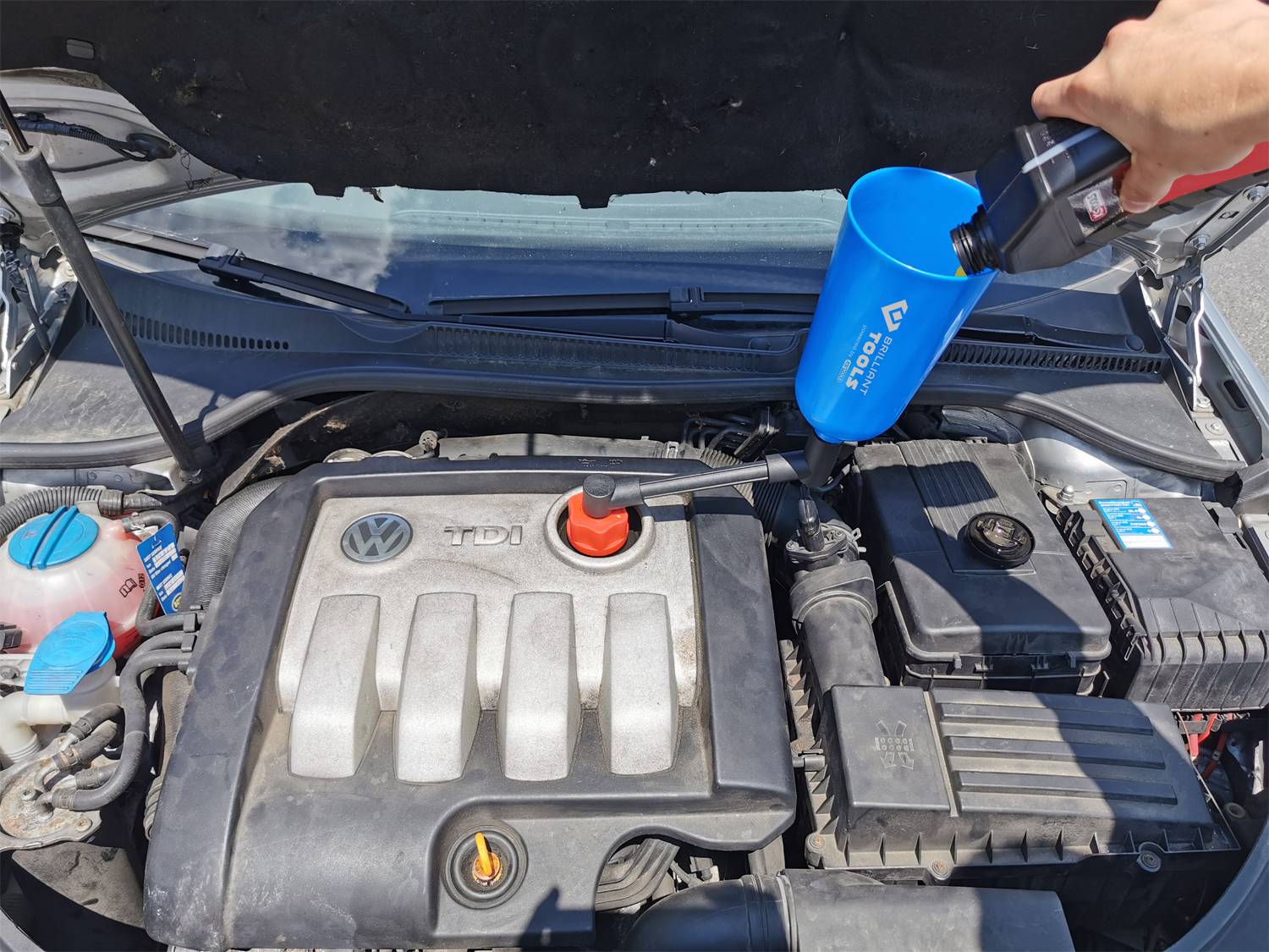 A person is pouring motor oil into a funnel positioned in the engine bay of a car. The bonnet is open.