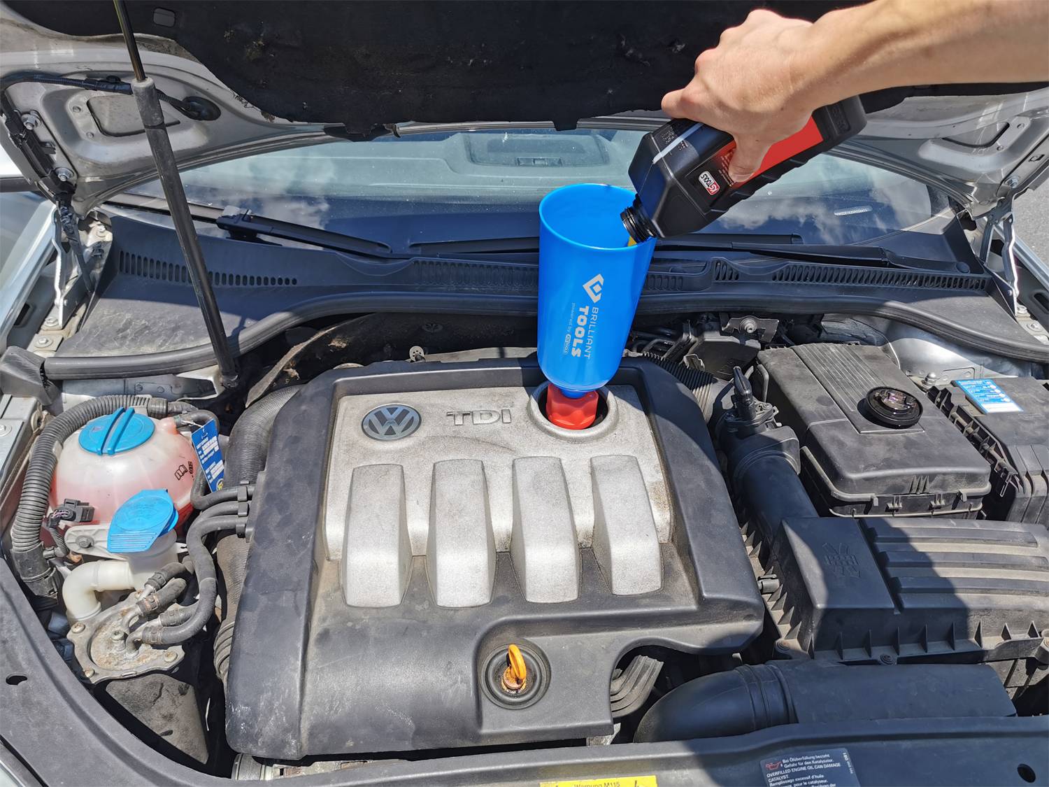 A person is topping up the oil in a car's engine with the bonnet open. A funnel is being used to pour the oil cleanly.