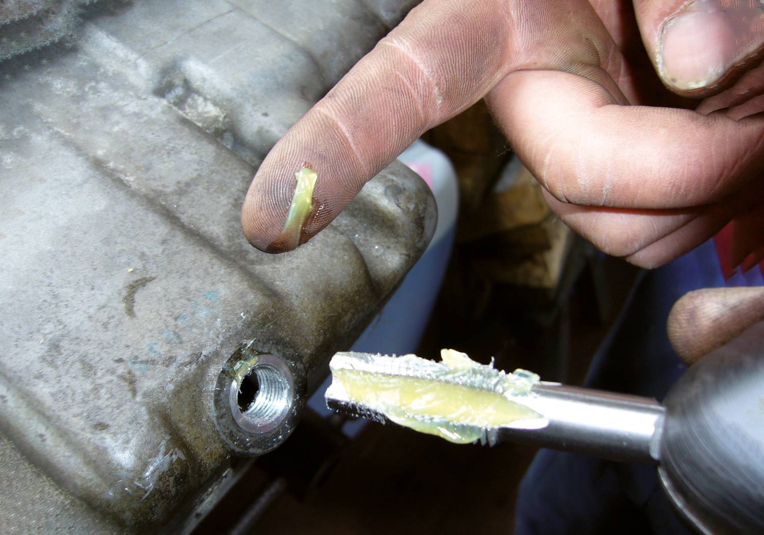A person is pointing at a drill with a drill bit covered in metal shavings and lubricant, close to a metal surface.