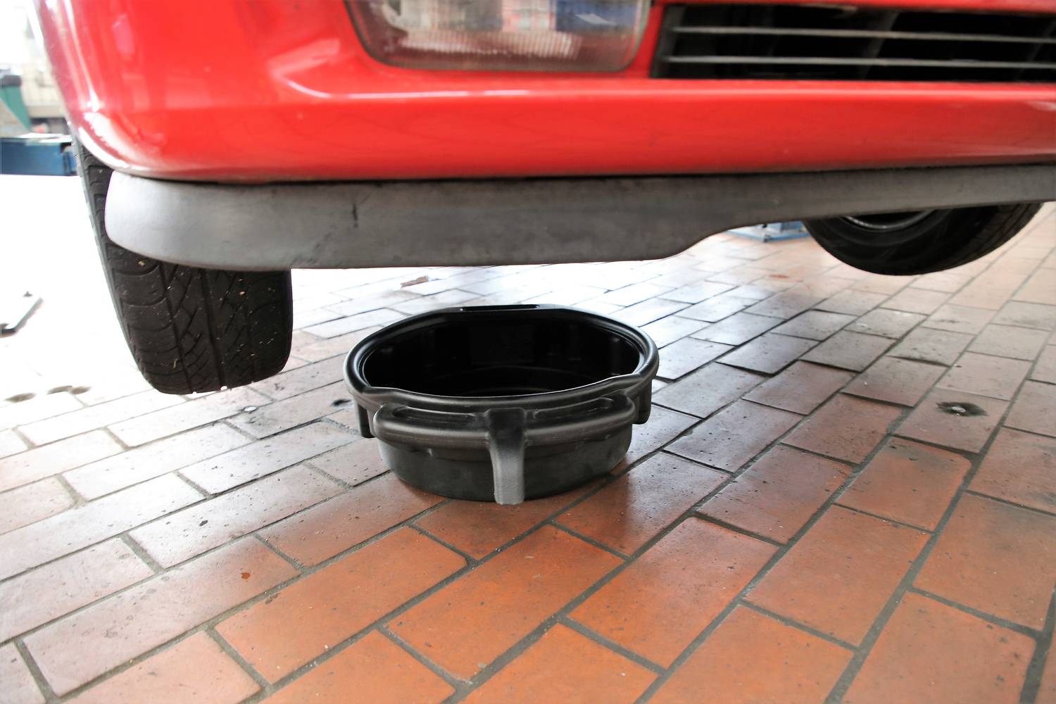 A black oil drip tray is positioned beneath a red car. The car is located in a garage with a tiled floor.