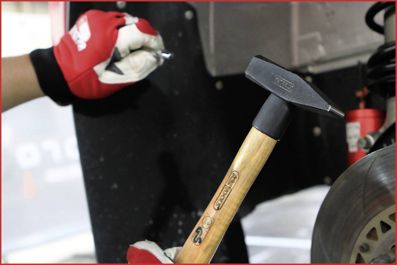 A person wearing red gloves is holding a small metal object, while another hand is holding a hammer next to a brake disc.