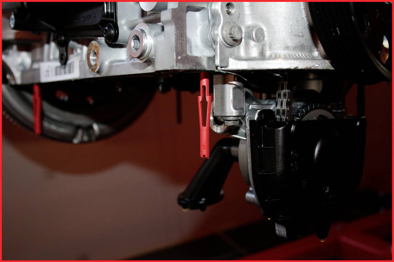 'Close-up of an engine component with timing belt and metal parts. A red plastic clip is visible. Technical detailed image.'