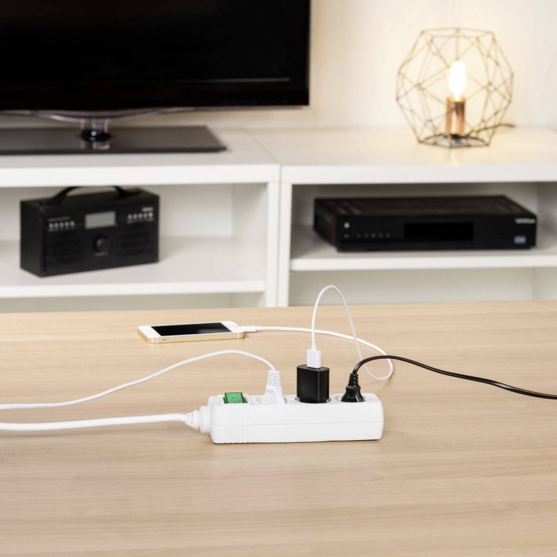 A table with a power strip, with two devices being charged. In the background, there is a radio, a television, and a lamp.