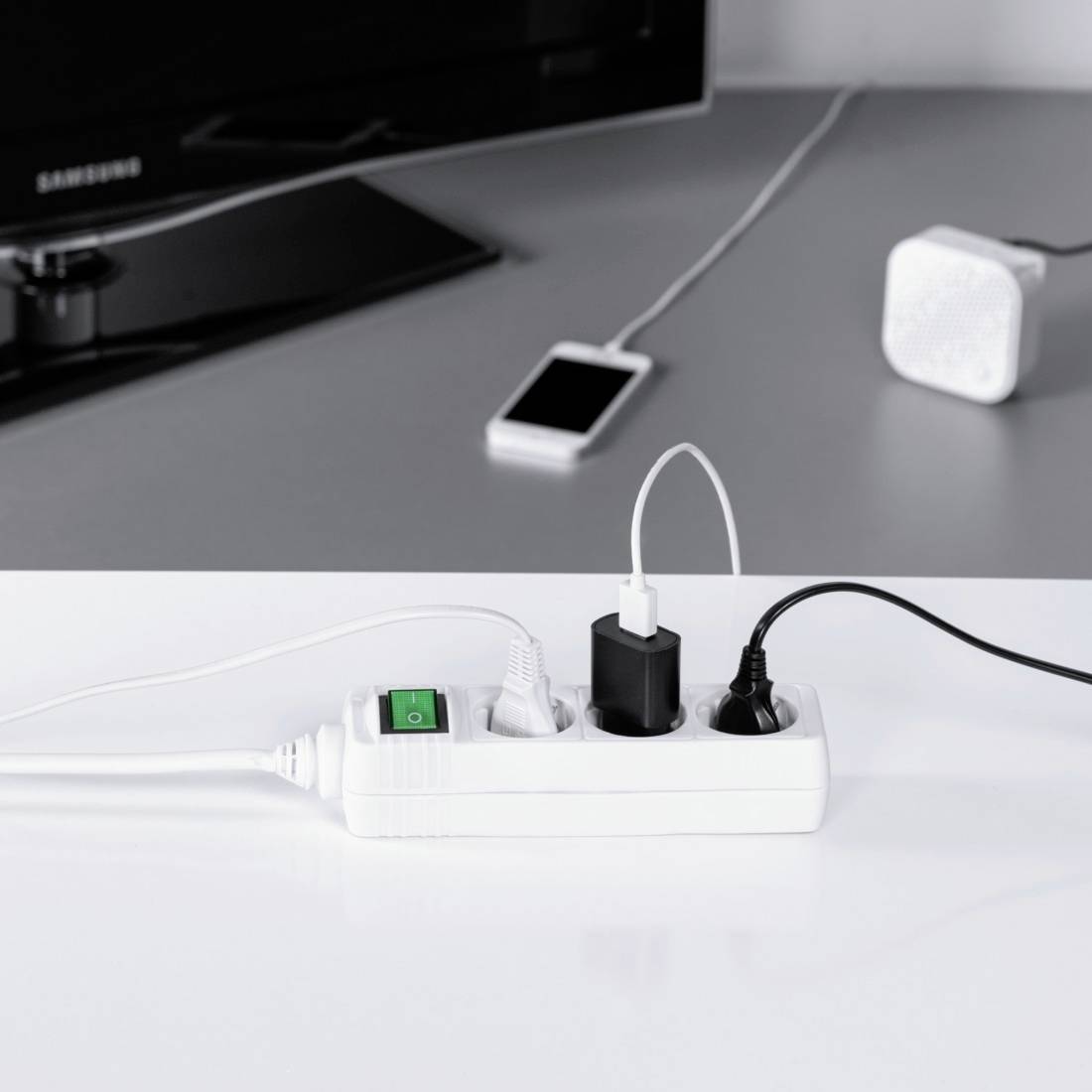 A white multi-socket adaptor with a green switch powers a smartphone charger and two other devices in front of a dark screen.