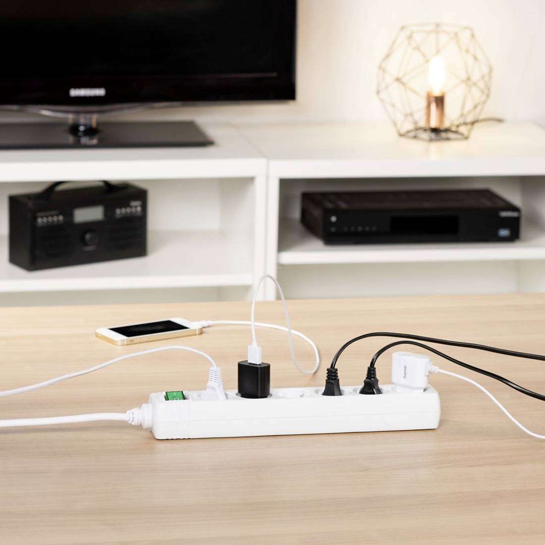 A multi-socket extension lead on a table. A smartphone charging via a USB charger, surrounded by electronic devices in the background.