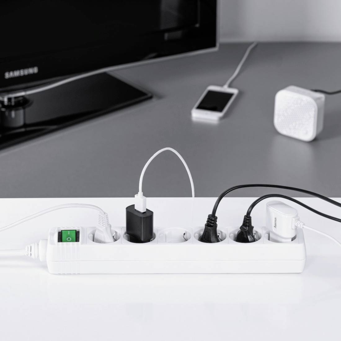 A white multi-socket extension lead with its switch turned on, to which several devices, including a mobile phone and a speaker, are connected.