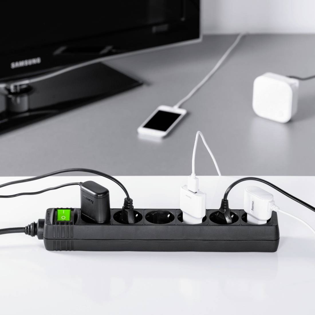 A six-socket extension lead, with four sockets in use. A television and a smartphone are visible in the background.