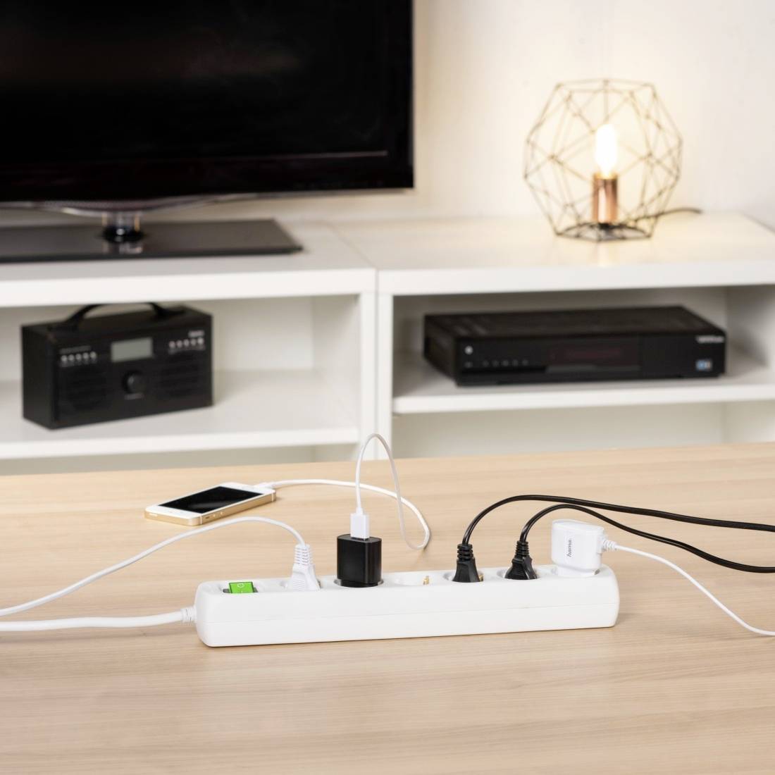 A table with a multi-socket extension lead, to which several devices are connected. A television and a lamp are visible in the background.