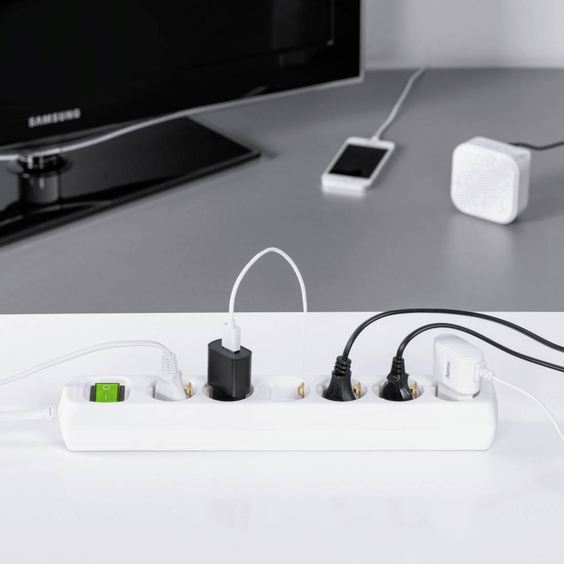A multi-socket extension lead on a table supplies electronic devices such as a smartphone and speakers. A television is positioned in the background.