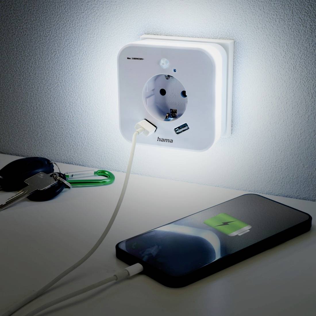 A mobile phone is being charged on a table. It is plugged into an illuminated socket in the wall, next to a bunch of keys.