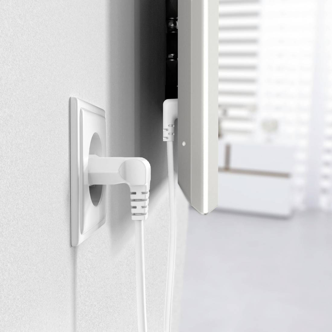 Power socket with a white plug on the wall, connected by a cable to a device. Blurred interior in the background.
