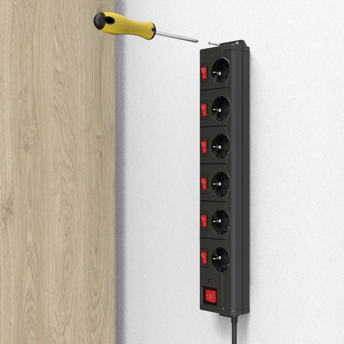 A black power strip with switches is mounted on a white wall. A screwdriver is inserted at the top of the strip.