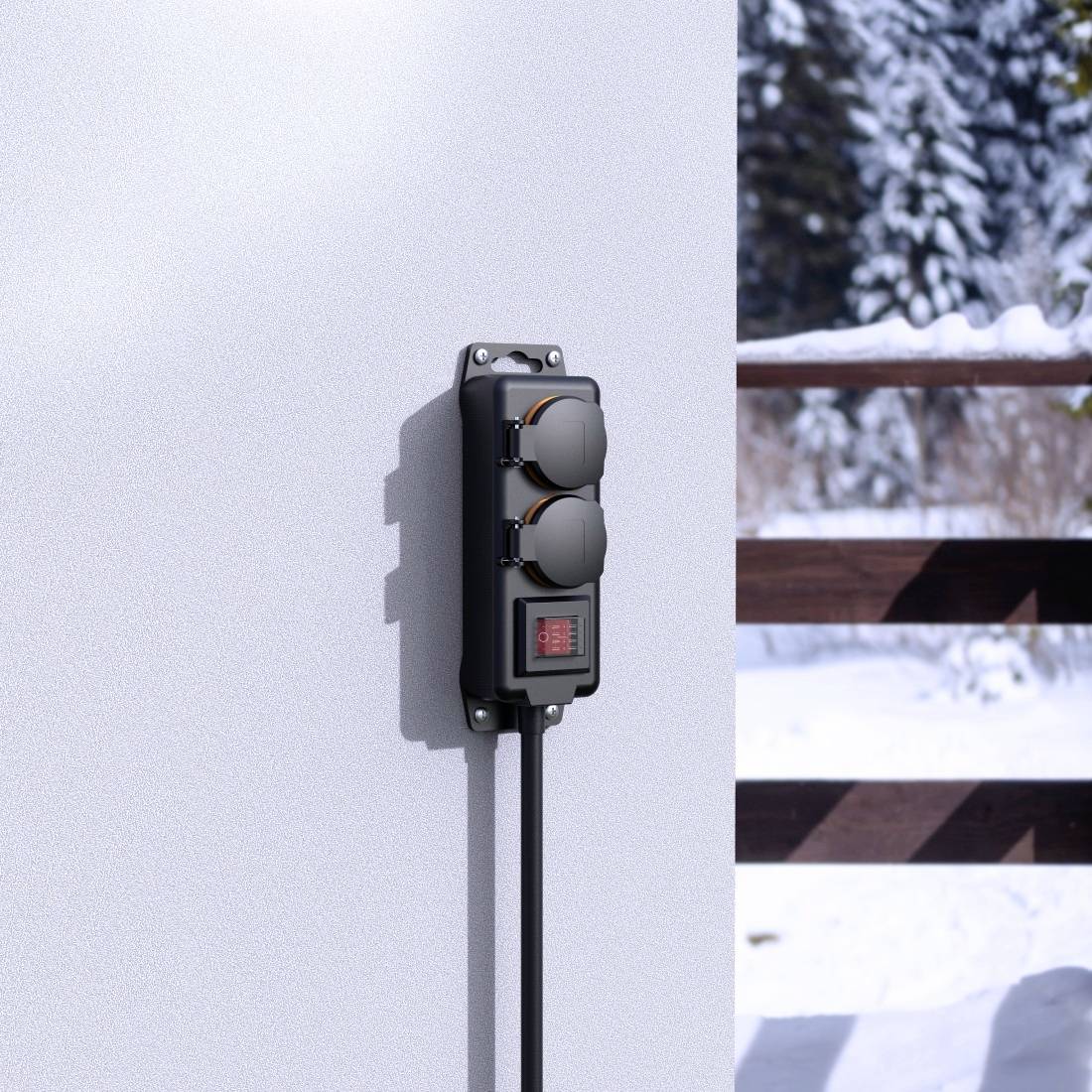 A weatherproof outdoor socket is mounted on a white wall. In the background, snow-covered trees and a wooden fence are visible.