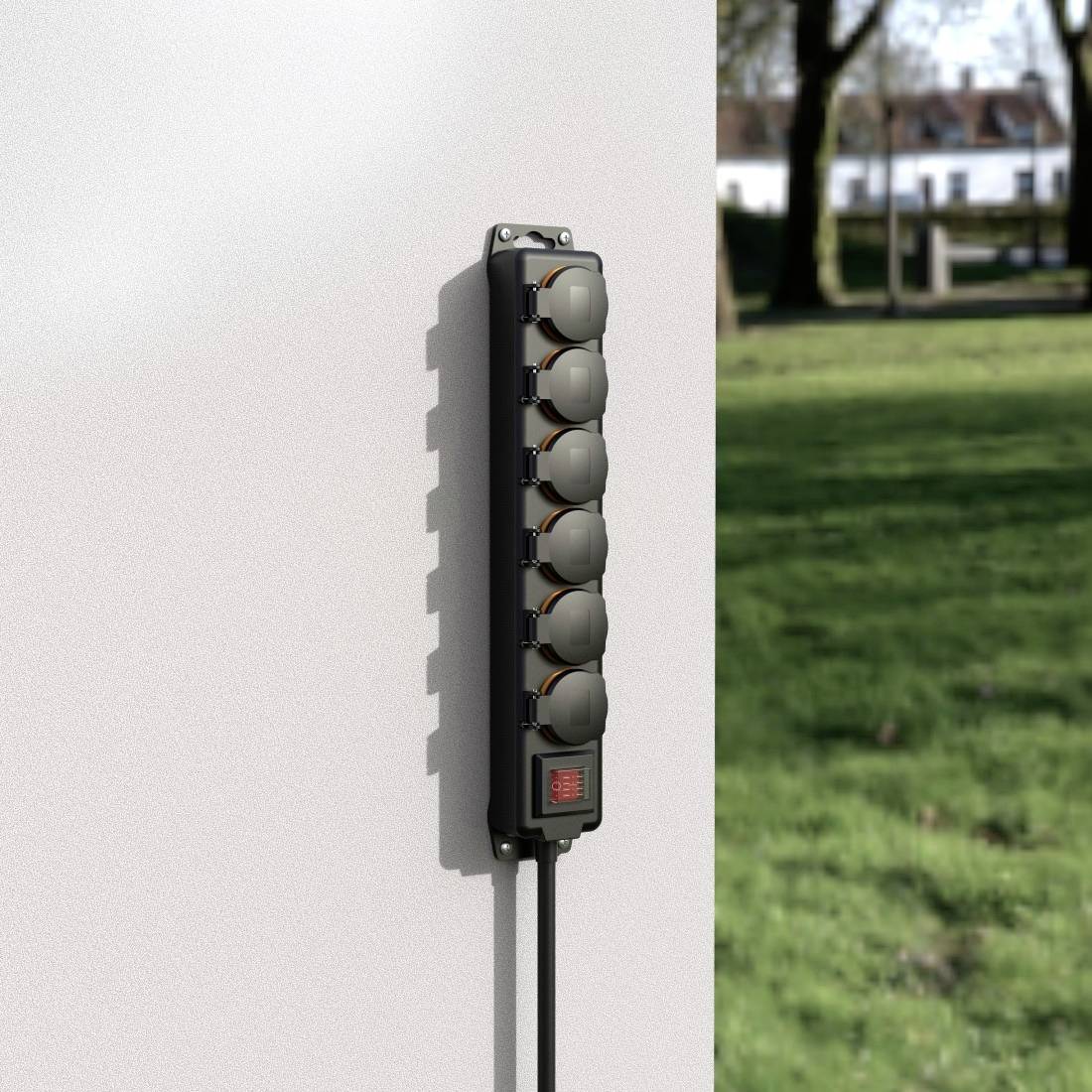 A vertical six-socket power strip is mounted on a wall outdoors. Grass is visible in the background.