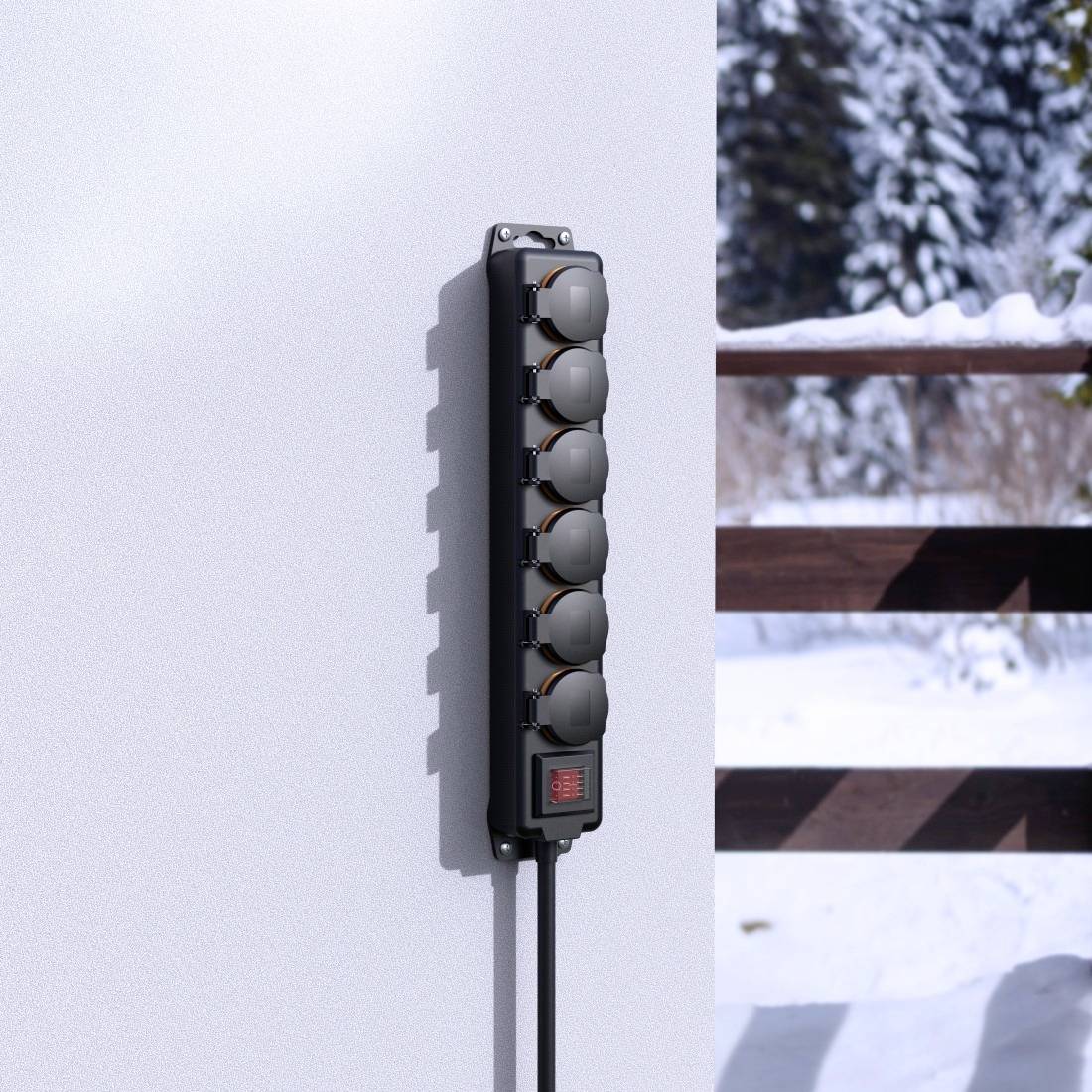 A black six-socket extension lead mounted on a wall, with a snowy landscape and wooden fence in the background.