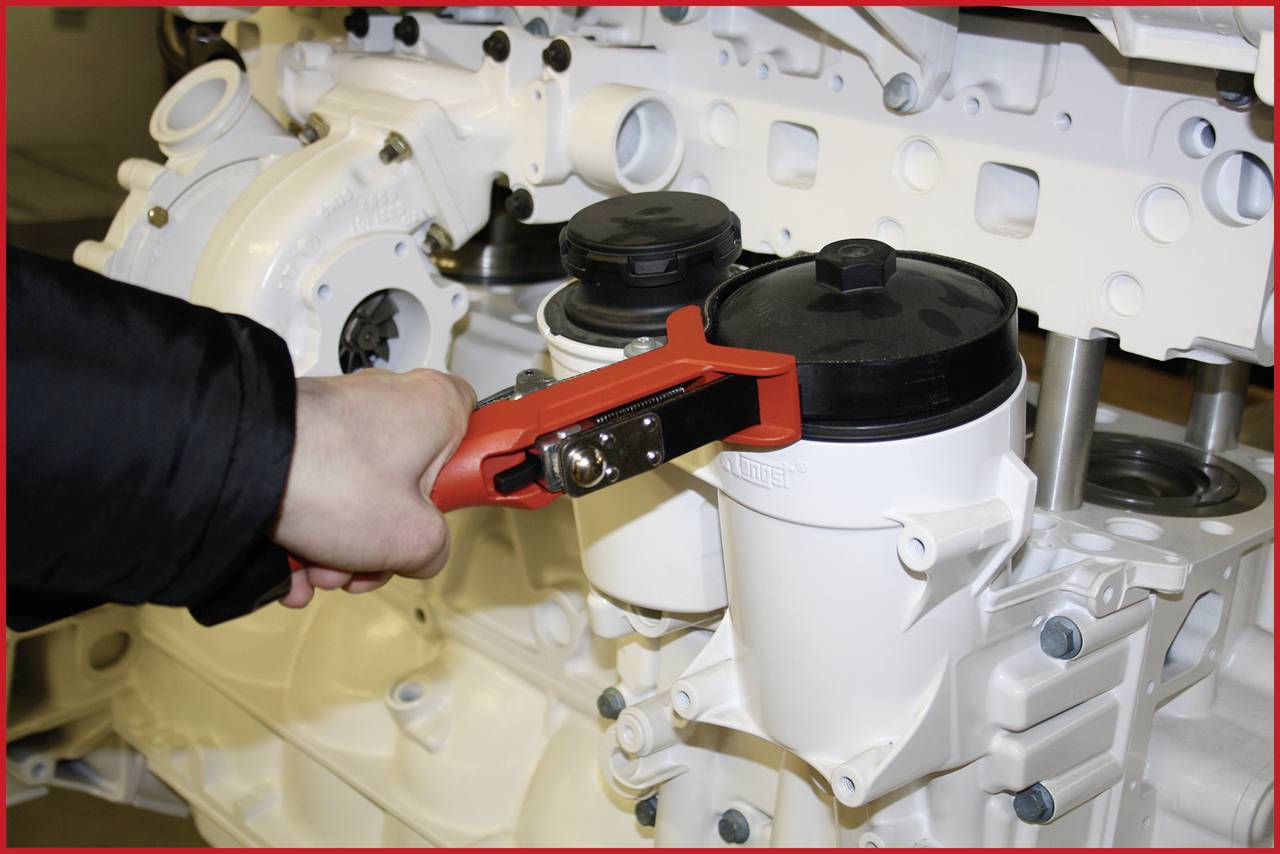 A person is using a red tool to turn or open the black lid of a white filter on a large machine.