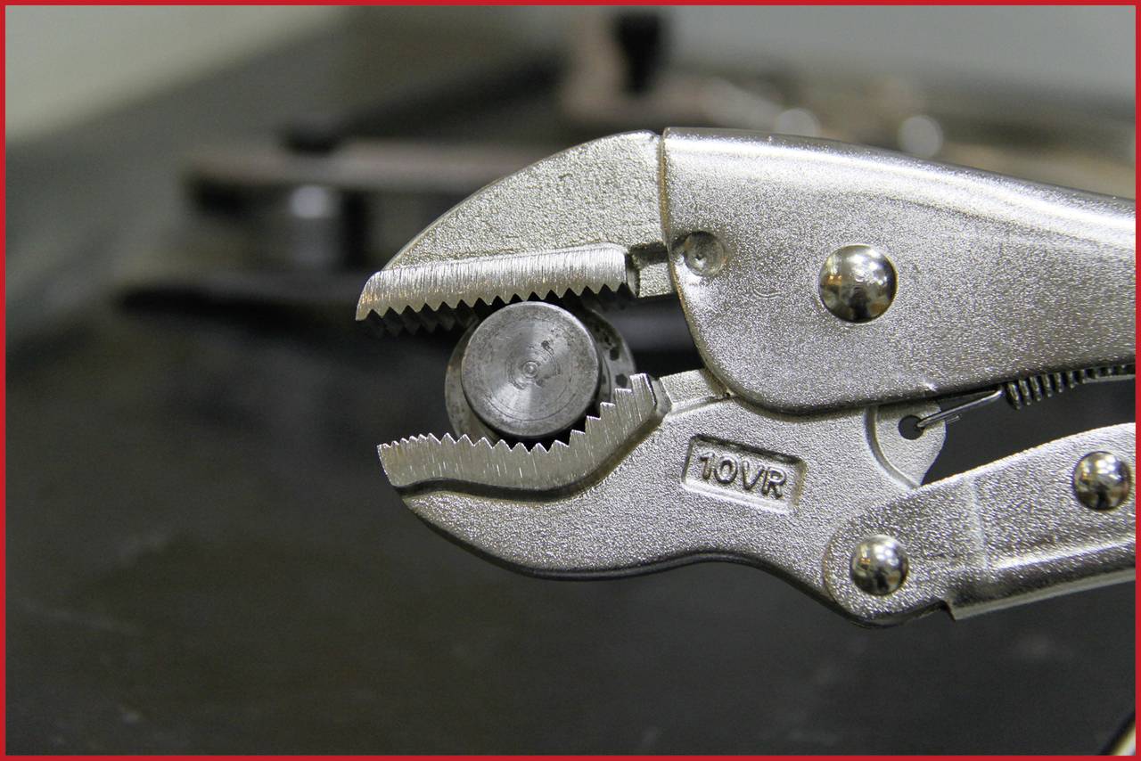 A silver pair of pliers grips a small metal nut. Blurred tools can be seen in the background.