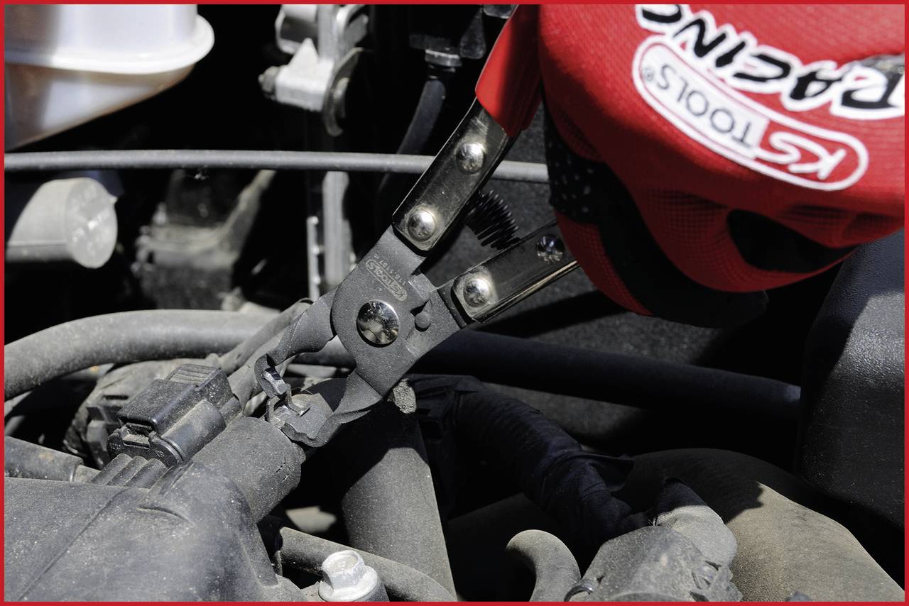 A hand wearing a red work glove is using pliers to work on a car part. Several cables and hoses are visible.