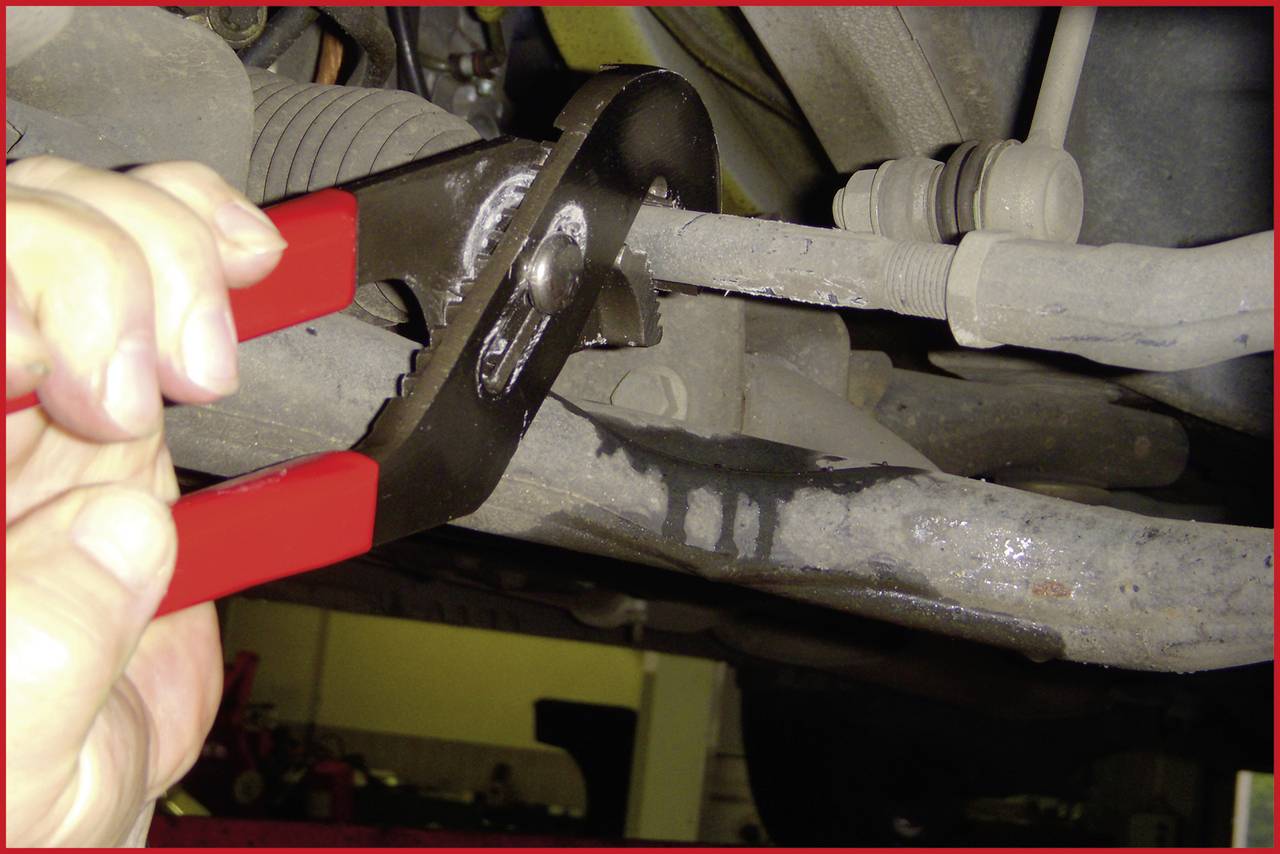 A hand holds a pair of pliers and repairs a part of a car. In the background, car parts and a workshop are visible.