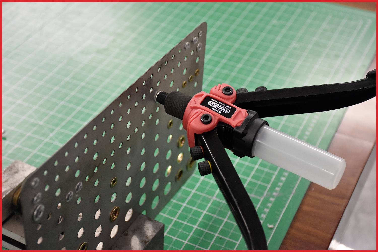 A riveting pliers sets rivets into a metal plate with holes, which is lying on a green cutting mat.