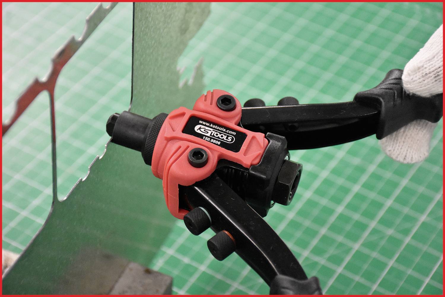 A person is cutting a piece of sheet metal with a red tin snips against a green mesh background.