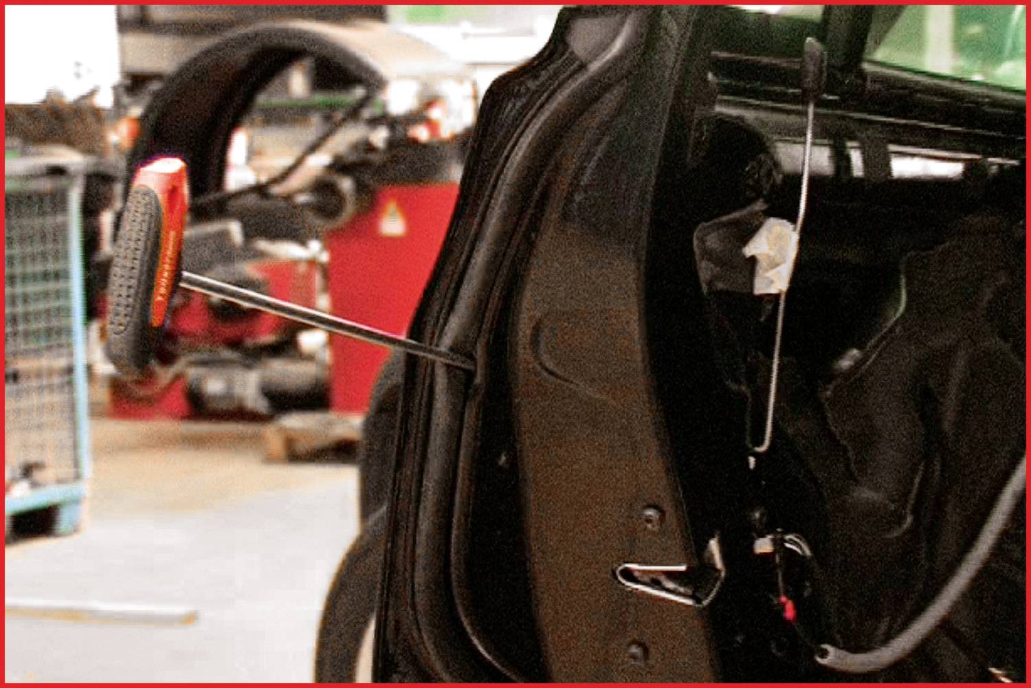 A car is being worked on by a mechanical device that extends through the open car door. Workshop equipment is visible in the background.