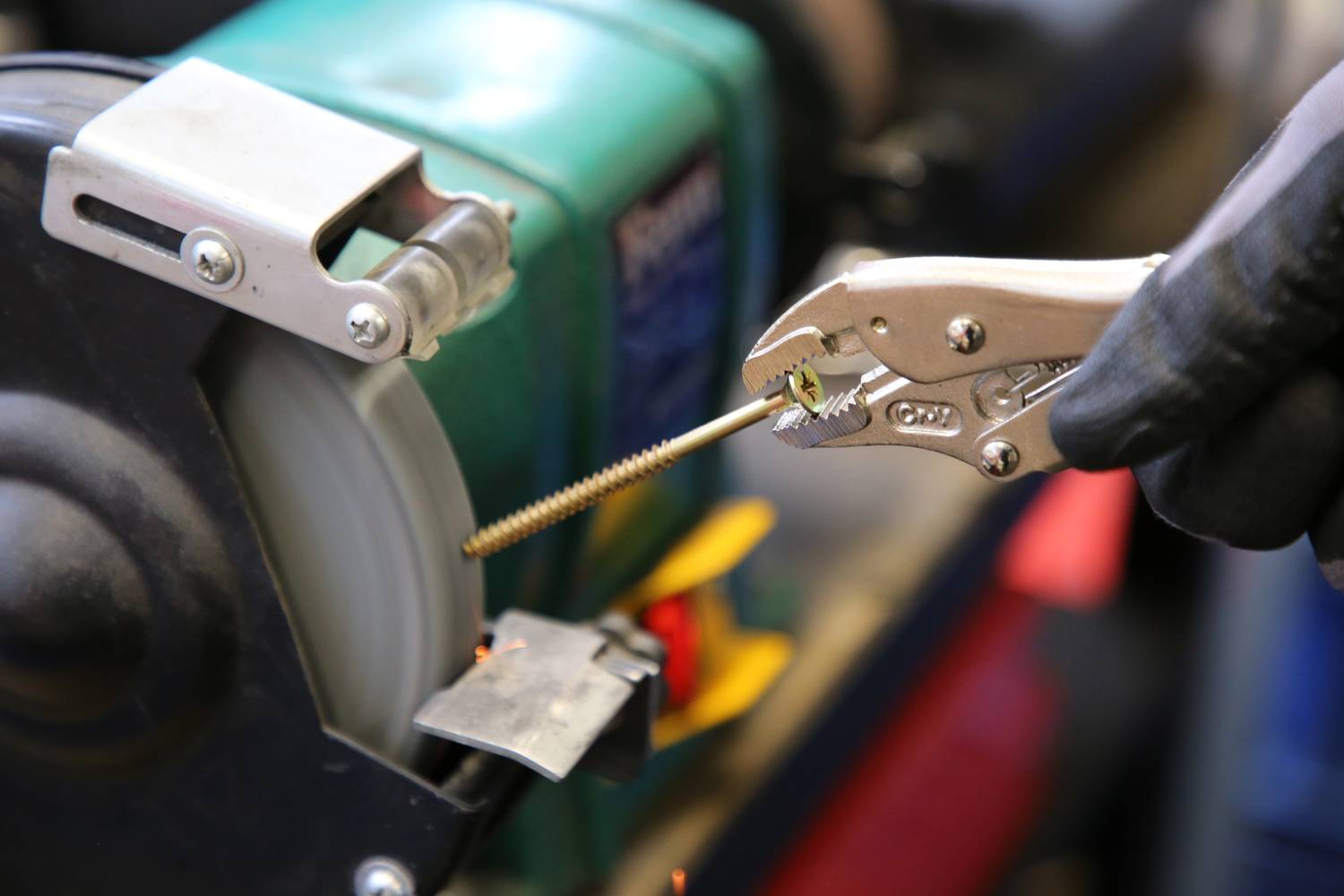 A screw is being sharpened on a grinding machine. A spanner holds the screw firmly, while someone wearing a glove performs the work.