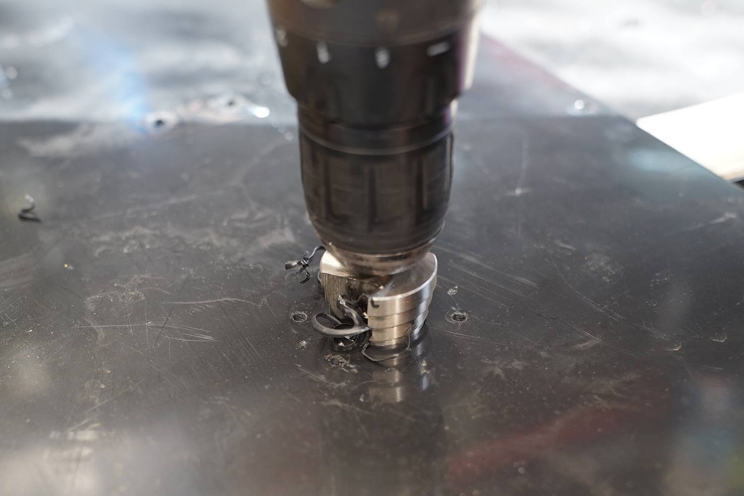 A drill is boring a hole into a black metal piece, with metal shavings surrounding the hole.