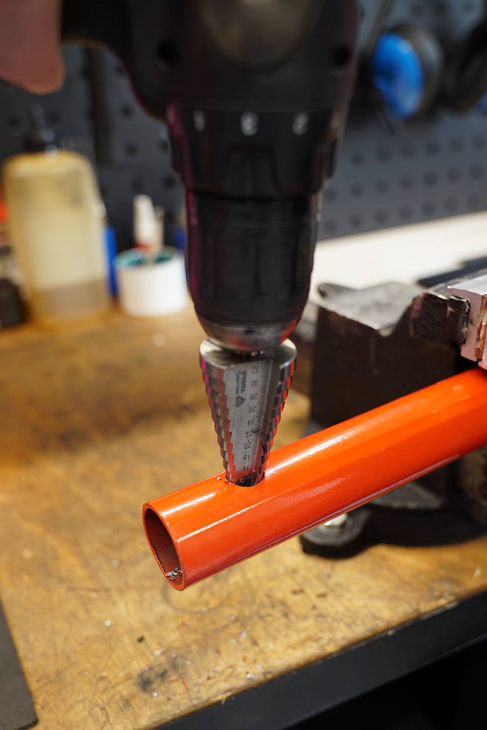 A drill with a cone drill bit is creating a hole in an orange metal pipe on a workbench in a workshop.