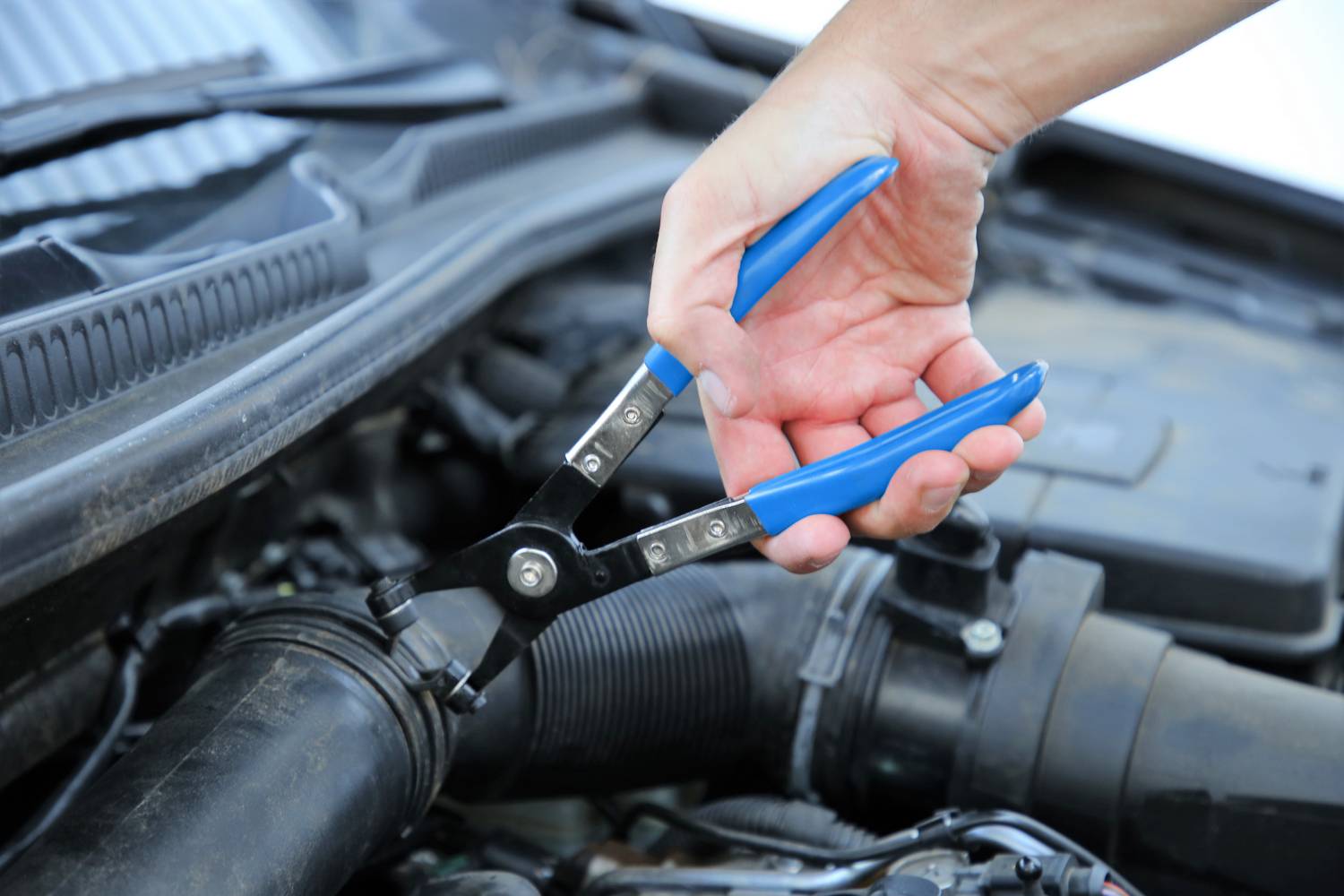 A hand holds pliers over the open engine compartment of a car. The pliers are being used to tighten a hose clip on an engine line.
