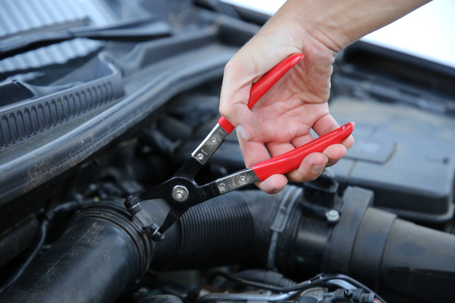 A hand is using a red pair of pliers to work on a car. The focus is on the pliers and the engine compartment.