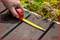 A hand holds a tape measure on a wooden decking next to colourful plants, measuring the length of the decking boards.