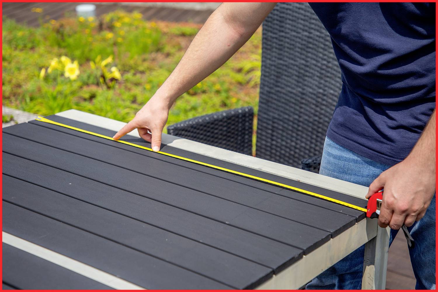 A person is measuring the width of a black wooden table outdoors using a measuring tape. Plants are visible in the background.